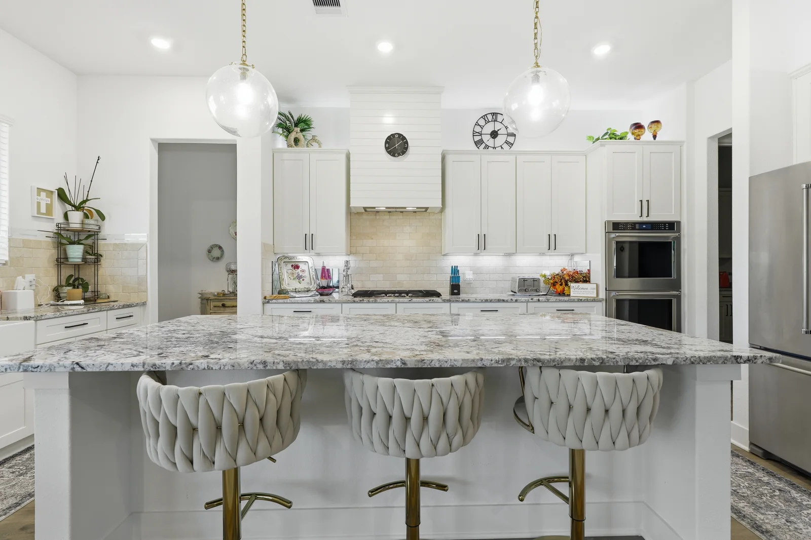 White kitchen with a patterned stone island, three upholstered stools, and glass pendant lights.